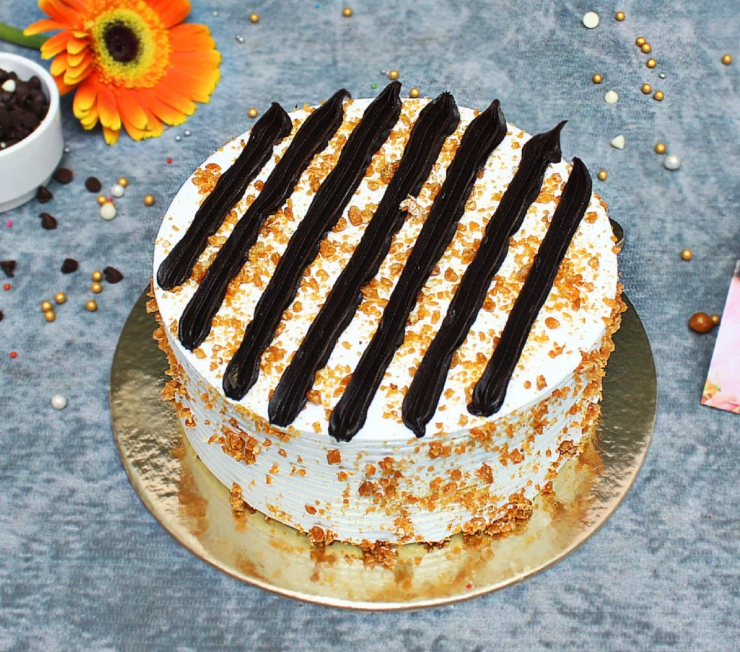 A frosted cake topped with dark chocolate stripes and coated around the sides with crunchy butterscotch nuts near me, displayed on a golden base.